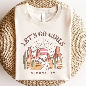 May include: A white t-shirt with a graphic print of a desert landscape with the text "Let's Go Girls Sedona, AZ".