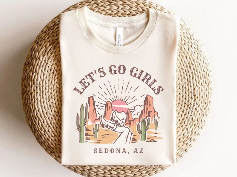 May include: A white t-shirt with a graphic print of a desert landscape with the text "Let's Go Girls Sedona, AZ".