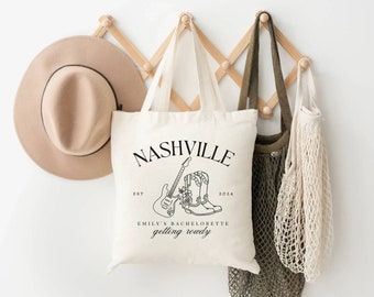 以下が含まれることがあります： 黒い文字で「Nashville Est. 2024 Emily's Bachelorette Getting Rowdy」と書かれた白いトートバッグ。ギターとカウボーイブーツに花が描かれた黒い線画が描かれている。