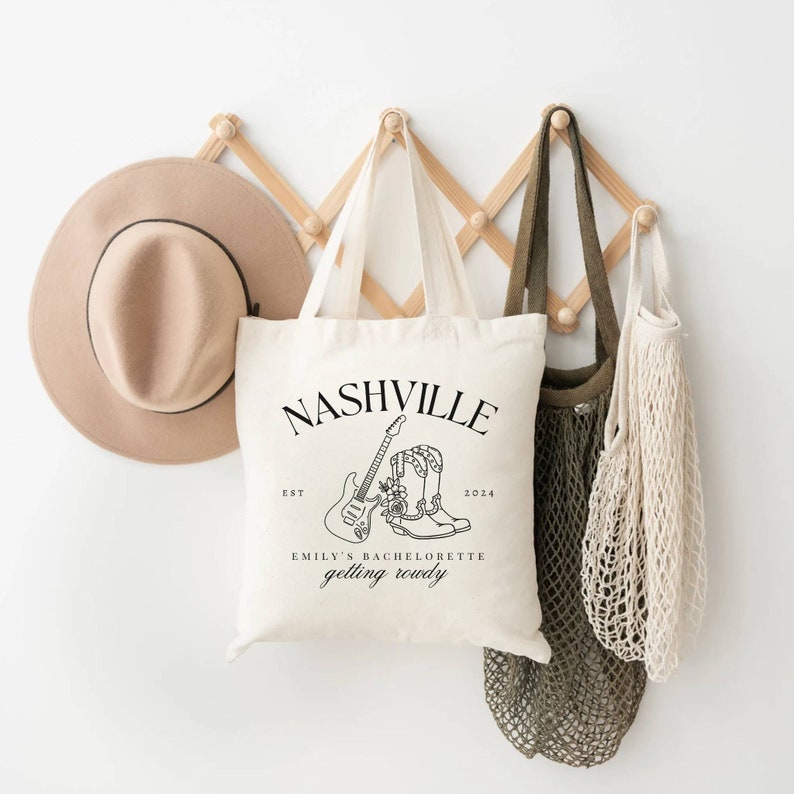 May include: A white tote bag with black text that reads "Nashville Est. 2024 Emily's Bachelorette Getting Rowdy". The bag has a black line drawing of a guitar and cowboy boots with flowers.