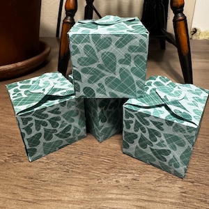 May include: Four small, square gift boxes with a green and white heart pattern. The boxes are arranged on a wooden surface, with a dark wooden chair and a potted plant in the background. The boxes are closed and ready for gifting.