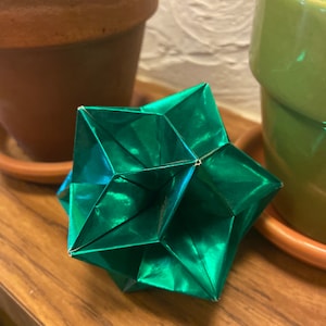 Pode incluir: Uma estrela de origami verde com um brilho metálico. A estrela é dobrada de uma única folha de papel e tem um design geométrico.