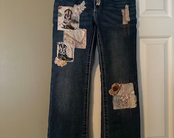 May include: Blue denim jeans with embroidered patches of cowboy boots and hats. The jeans have a bootcut style and are labeled "Wrangler Willow".