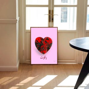 Può includere: Una stampa artistica incorniciata con un design a forma di cuore in rosso e nero su sfondo rosa. L'opera d'arte include testo arabo. La stampa è esposta in una stanza con pavimento in legno e una porta.