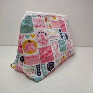 May include: A zippered pouch featuring a white background with a colorful pattern of pink ribbons, hearts, and inspirational text including "Think Pink," "You Are A Warrior," "Brave & Strong," "Support Admire Remember," and "Never Give Up."