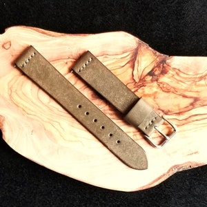 May include: A two-part watch strap in olive green. The leather strap has visible stitching and a silver buckle. The strap is displayed on a wooden surface, set against a dark background.