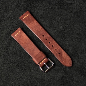 May include: Two-part brown leather watch strap with orange stitching. The strap features a silver buckle and several adjustment holes. The leather has a distressed look, and the strap is displayed against a dark background.