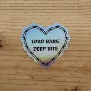 May include: A heart-shaped holographic sticker featuring a black barbed wire border and the text LOUD BARK DEEP BITE printed in a bold, dark font in the center. The sticker has a reflective, iridescent finish that shifts colors across its surface.