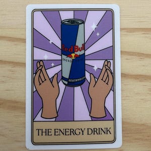 May include: A rectangular sticker featuring a Red Bull energy drink can in the center, set against a purple and white striped background. Two hands are positioned on either side of the can. The text "THE ENERGY DRINK" is at the bottom.