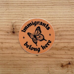 May include: A circular sticker with a matte orange background featuring a black butterfly illustration in the center. The text "Immigrants belong here" is printed in a black serif font, curving around the butterfly with two small stars.