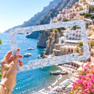 May include: White license plate frame with a blue seashell pattern. The frame has a rectangular shape with rounded corners and pre-drilled holes for mounting. The design includes various sea creatures.