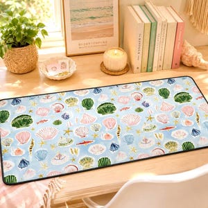May include: A rectangular desk mat featuring a light blue background with a repeating pattern of various seashells, starfish, and small orange fish. The mat has a black stitched border and a smooth surface, displaying a variety of marine life.