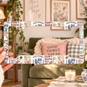 May include: A white license plate frame featuring a repeating pattern of illustrations including a raccoon, a duck, and a llama, with the text "DON'T DUCK WITH ME," "STAY TRASHY," and "MESSY HAIR" printed in a dark blue and red color scheme.