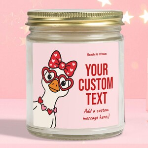 Può includere: Una candela in barattolo di vetro con coperchio dorato ed etichetta rosa con un'oca dei cartoni animati con occhiali a forma di cuore e un fiocco. L'etichetta dice "YOUR CUSTOM TEXT" in rosso. Lo sfondo è rosa con stelle.