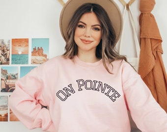 Puede incluir: Una sudadera rosa claro con el texto "ON POINTE" en letras negras arqueadas. La sudadera tiene cuello redondo y mangas largas. La modelo lleva vaqueros azules y un sombrero beige.