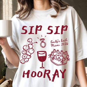 May include: White t-shirt with a crimson design. The design includes the words "SIP SIP HOORAY", a ring, grapes, a wine glass, and a cake. Text on the shirt reads "Emily's bach Miami 2026".