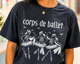 May include: A dark charcoal gray short-sleeve t-shirt featuring a white graphic of three skeletons wearing ballet tutus and hair bows. The text "corps de ballet" is printed in white above the skeletons, accompanied by small white bat silhouettes.
