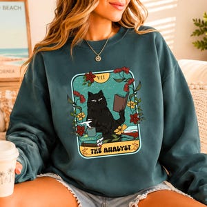 Puede incluir: Una sudadera de cuello redondo color verde bosque con un estampado gráfico de un gato negro leyendo un libro y sosteniendo una taza. El diseño está enmarcado como una carta del tarot con flores y el texto VII THE ANALYST. Mangas largas.