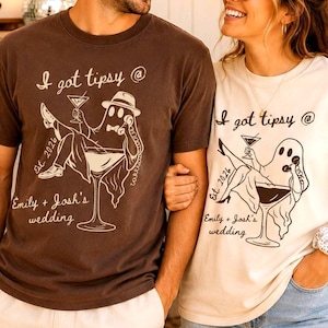 May include: A dark brown t-shirt and a cream t-shirt, each featuring a black graphic of a ghost sitting in a martini glass, holding a telephone, with the text I got tipsy @, Est. 2026, and Emily + Josh's wedding printed in a script font.