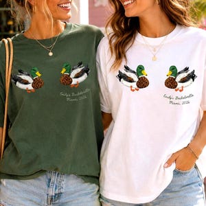 May include: Two short-sleeve crew neck t-shirts, one in olive green and one in white, each featuring a graphic of two mallard ducks and the text "Emily's Bachelorette Miami, 2026" printed on the left chest area.