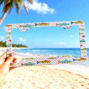 May include: A rectangular license plate frame featuring a white background with a black polka dot pattern and a repeating design of colorful fish in shades of blue, green, yellow, and orange, including four mounting holes for attachment.
