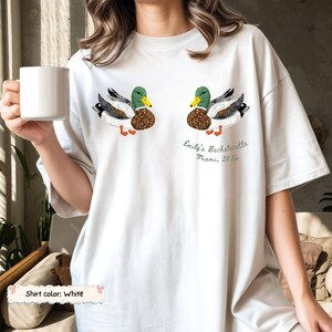 May include: A white t-shirt featuring two mallard duck illustrations with patterned wings and bodies. Below the right duck, the text reads: Emily's Bachelorette Miami, 2026. The shirt has short sleeves and a crew neckline, presented in a relaxed fit.