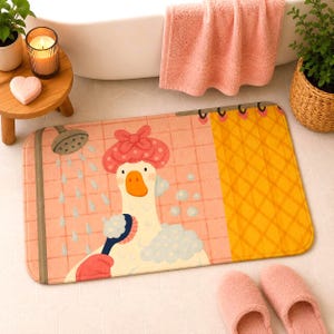 May include: A rectangular bath mat featuring a cartoon goose wearing a pink polka-dot shower cap, holding a blue scrub brush with white soap suds. The background shows a pink tiled shower wall with a shower head and a yellow curtain section.