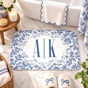 May include: A rectangular bathroom rug featuring a white center with the blue monogram letters A and K separated by a vertical line. The rug has a blue and white floral pattern border with intricate lace-like details around the edges.