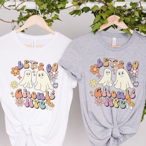 May include: Two white t-shirts with a retro-style graphic design featuring two ghosts, flowers, and the text "Let's Go Ghouls NYC".