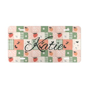 Puede incluir: Placa de matrícula rectangular con un diseño de patchwork en rosa, verde y melocotón. El patrón incluye fresas, flores y corazones. El nombre "Katie" está escrito en cursiva en el centro.