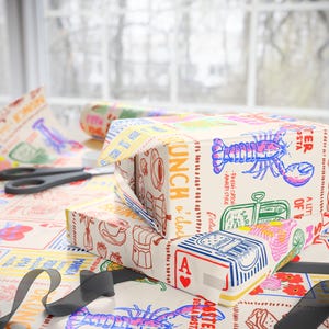May include: A gift box wrapped in colorful paper with a lobster illustration and the words "Lunch o'clock". The wrapping paper features various food-related graphics and text, with a black ribbon and scissors nearby.