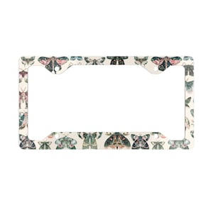 May include: Cream-coloured license plate frame featuring a repeating pattern of colourful moths. The moths are in shades of pink, green, blue, and black. The frame is rectangular with rounded corners and mounting holes.