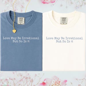 May include: Two Comfort Colors t-shirts, one dusty blue and one off-white, with the text "Love May Be Irrational But So Is π" printed on the front. A gold heart pendant necklace is draped over the blue shirt. Floral accents.