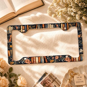 May include: A rectangular license plate frame featuring a dark blue background decorated with a repeating pattern of colorful book spines, delicate floral illustrations, and green leaves. The frame has four mounting holes and a smooth finish.