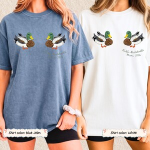 May include: A blue jean colored t-shirt and a white t-shirt, each featuring a graphic of two mallard ducks. The white shirt includes the text 'Emily's Bachelorette Miami, 2026' printed below the ducks. Both shirts have a crew neck and short sleeves.
