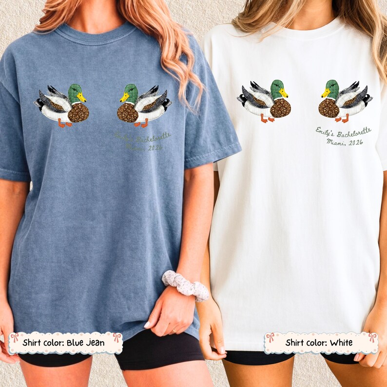 May include: A blue jean colored t-shirt and a white t-shirt, each featuring a graphic of two mallard ducks. The white shirt includes the text 'Emily's Bachelorette Miami, 2026' printed below the ducks. Both shirts have a crew neck and short sleeves.