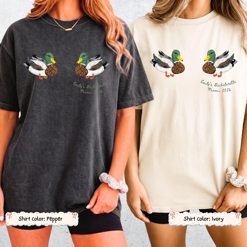May include: Two short-sleeve crewneck t-shirts, one in pepper gray and one in ivory, each featuring a graphic of two mallard ducks with green heads and patterned bodies. Below the ducks, the text reads Emily's Bachelorette Miami 2026.