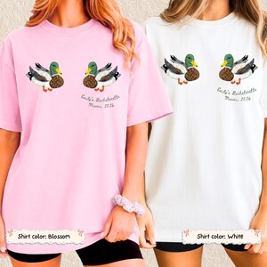May include: A light pink t-shirt and a white t-shirt, each featuring two mallard duck illustrations and the text "Emily's Bachelorette Miami, 2026" printed in black. The ducks have green heads, brown chests, and white bodies with black wing details.