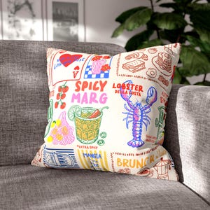 May include: A decorative throw pillow with a cream background and colorful illustrations. The pillow features images of food, cocktails, and text including "Spicy Marg" and "Lobster Della Costa". The design includes a lobster, tomatoes, and a cocktail.