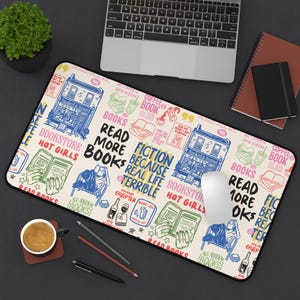 Könnte beinhalten: Ein rechteckiges, beiges Mauspad mit einem Design zum Thema Bücher. Das Design umfasst Illustrationen eines Buchladens, Bücher, Kaffeetassen und Text, der "Read More Books" und "Fiction Because Real Life is Terrible" lautet.