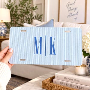 May include: A rectangular license plate featuring a light blue and white pinstripe pattern. The center displays the blue capital letters M and K separated by a vertical line. The plate has four mounting slots, one in each corner.