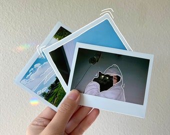 May include: A person holding a Polaroid photo of themself with a black cat. The photo is in a white frame with a blue sky background.