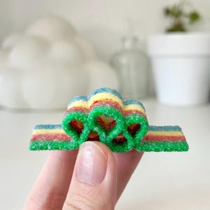 May include: A colorful gummy candy with a rainbow of colors, including green, blue, yellow, and red. The candy is shaped into a unique design, with a sugared coating. The candy is held in a hand.