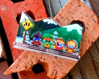 May include: A clear acrylic standee featuring four cartoon characters in winter gear standing in a snowy mountain landscape. A yellow diamond-shaped sign with a black graphic of two children walking is visible on the left side of the background.