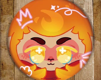 May include: A circular pinback button featuring a stylized orange character with large, glowing yellow eyes, dark eyebrows, and a small nose. The design includes a pink crown outline, white sparkles, and pink abstract shapes on an orange background.