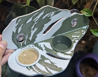 May include: A white and green 3D printed leaf-shaped feeder with two small bowls. One bowl contains a light brown substance and the other bowl is empty. The feeder has two suction cups on the back.