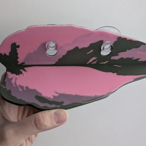 May include: A pink and green leaf-shaped object with two clear suction cups. The object has a pink base with green and darker pink accents. The object is held by a hand against a white background.