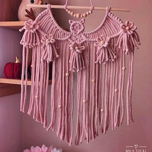 Pode incluir: Uma tape&ccedil;aria de parede de macram&eacute; rosa com uma haste de madeira e detalhes de contas. A pe&ccedil;a feita &agrave; m&atilde;o apresenta um design de coruja e enfeites florais. Fios verticais pendem por baixo, com contas de madeira. O texto "POLY TUSAL Handmade" &eacute; vis&iacute;vel.