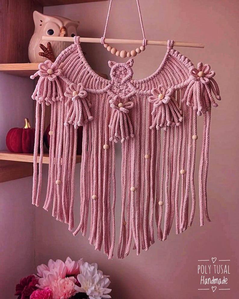 Pode incluir: Uma tape&ccedil;aria de parede de macram&eacute; rosa com uma haste de madeira e detalhes de contas. A pe&ccedil;a feita &agrave; m&atilde;o apresenta um design de coruja e enfeites florais. Fios verticais pendem por baixo, com contas de madeira. O texto "POLY TUSAL Handmade" &eacute; vis&iacute;vel.