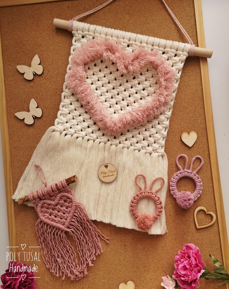 Pink macrame rabbits pendants with thin ears at cork board, next to Macrame wall Hanging with fluffy heart and Heart Hanger. Poly Tusal Handmade logo in the left down corner.